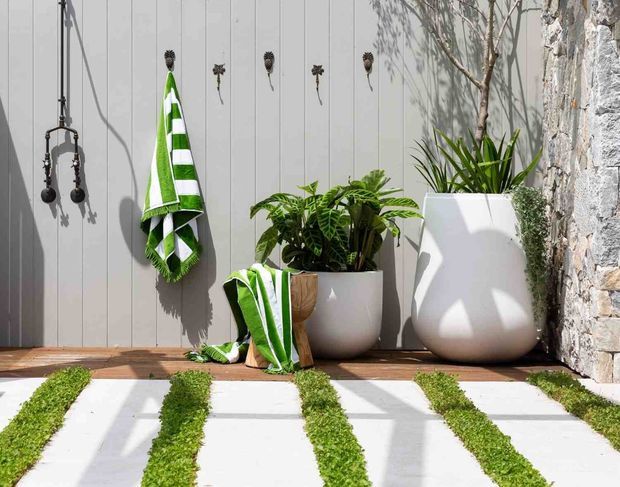 A bright landscaped courtyard or outdoor patio vignette with white vertical wall panelling, a timber ledge, hanging decorative bells, green-and-white striped towels, large white planters with leafy plants, and a pale stone wall. The foreground features light paving slabs separated by narrow strips of grass, creating a patterned garden path.
