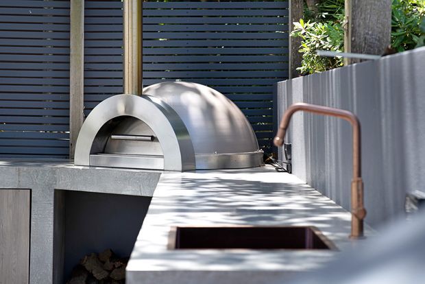 Close-up view of a built-in outdoor cooking area featuring a stainless-steel dome pizza oven set into a concrete benchtop. The installation includes a dark horizontal-slat privacy screen, grey corrugated metal side cladding, exposed copper-toned pipework or handrail, and a recessed opening in the benchtop. Dappled sunlight falls across the outdoor kitchen work surface.