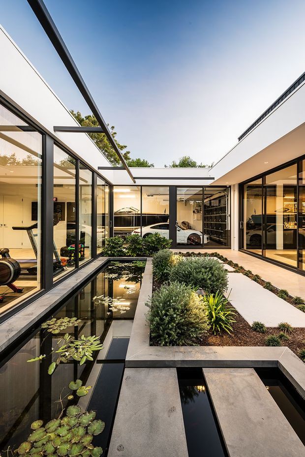 A contemporary courtyard viewed from within a modern residence, featuring a long reflective pond with lily pads, geometric concrete stepping slabs, and densely planted garden beds. Floor-to-ceiling black-framed glazing surrounds the courtyard, revealing bright interior spaces and a parked car beyond. White rendered walls, flat rooflines, and slim projecting steel elements create a clean, minimalist architectural composition.