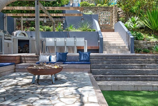 An outdoor entertaining area with a built-in stone and timber seating platform, blue cushions, a freestanding fire bowl, and a pergola with hanging lights. Stone-paved flooring, rendered and stone retaining walls, garden planting, an outdoor fireplace or wood storage nook, and steps leading to an upper terrace create a layered landscaped courtyard setting.
