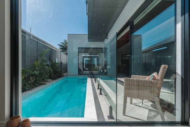 A narrow turquoise swimming pool runs alongside a modern glazed residence, viewed from a covered outdoor walkway or interior through floor-to-ceiling glass. The space features crisp white paving, dark metal-framed glazing, a covered ceiling, privacy fencing, tropical planting, and a woven outdoor lounge chair. Strong linear composition and reflections create a contemporary indoor–outdoor connection.