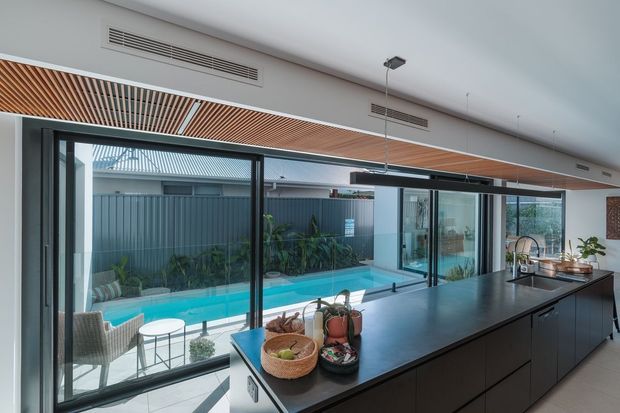 Contemporary open-plan kitchen with a long dark island, integrated appliances, timber slatted ceiling feature, and extensive floor-to-ceiling glazing overlooking a narrow swimming pool and enclosed outdoor area. The space has a clean, refined fit-out with strong indoor-outdoor connection.
