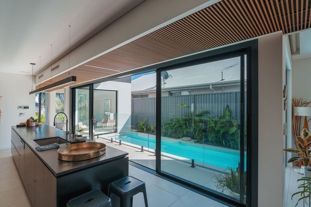 Bright contemporary kitchen opening directly onto a narrow swimming pool courtyard. The room features a long dark island with integrated sink and cooktop, pale cabinetry, large black-framed glazed sliding doors, light tiled flooring, and a timber slatted ceiling feature. Tropical planting and a high privacy fence are visible outside.