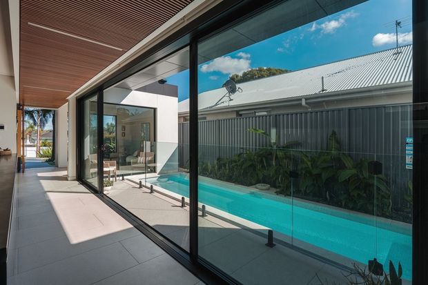 Contemporary indoor-outdoor corridor beside a long swimming pool, defined by extensive black-framed floor-to-ceiling glazing and sliding doors. The space has large light-grey tiled flooring, a warm timber slatted soffit, white rendered walls, and views toward a landscaped pool edge and neighbouring roofline. A dining area is visible through the glazing, while the main emphasis is the glazed circulation space and pool connection.