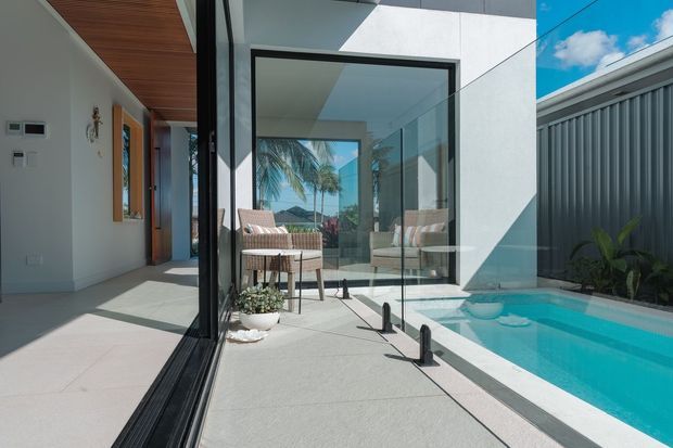 Bright contemporary residential courtyard viewed from an indoor-outdoor threshold, with large black-framed glazing, a covered terrace furnished with woven lounge chairs, pale tiled paving, and a narrow turquoise swimming pool beside a dark vertical-screen boundary fence. Warm timber soffit detailing, tropical planting, and palm trees are visible through the glazing, creating a strong indoor-outdoor connection.