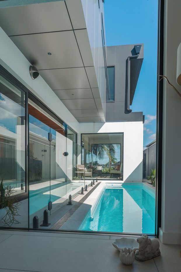 A bright contemporary courtyard-style residential space viewed through floor-to-ceiling glazing, with a narrow turquoise swimming pool enclosed by white rendered walls and modern glass doors. The architecture features clean rectilinear forms, a projecting upper level, an aluminium soffit with recessed lighting, and dark-framed glazing. Palm trees and blue sky are visible beyond the courtyard.