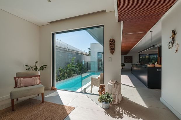 A bright contemporary open-plan interior connecting an entry or living area with a compact kitchen and a narrow turquoise swimming pool beyond full-height glass sliding doors. The space features pale tiled flooring, warm white walls, dark built-in kitchen cabinetry, a richly toned timber slatted ceiling, indoor plants, and a textured neutral rug with an upholstered accent chair. Strong indoor–outdoor connection and abundant natural light define the scene.
