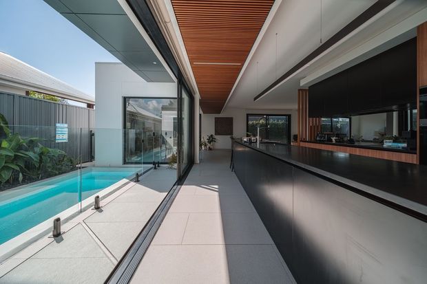 A contemporary open-plan interior with a long black kitchen or bar counter and integrated cabinetry on the right, floor-to-ceiling glazing on the left, and a partially covered outdoor swimming pool visible beyond the glass. The ceiling features warm timber slats, while large pale tiles and dark joinery create a refined indoor-outdoor connection.