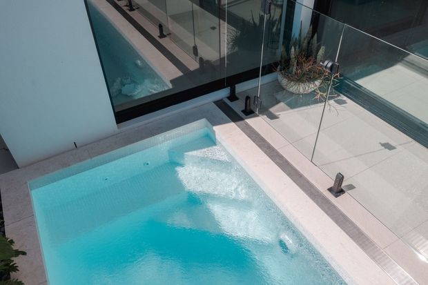 A modern outdoor swimming pool viewed from above, adjoining a pale tiled terrace and enclosed by clear glass balustrades. The pool features bright turquoise water, integrated steps, and visible water jets, with dark metal framing and minimal contemporary detailing.