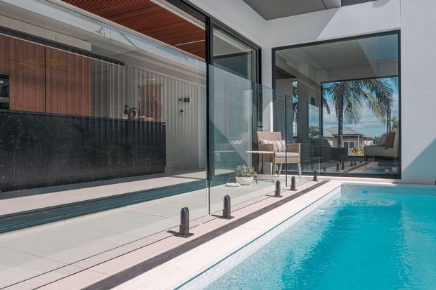 A bright contemporary indoor-outdoor pool courtyard with turquoise water beside large-format pale paving. Black-framed floor-to-ceiling glazing opens to a refined interior featuring a dark stone island or counter, warm timber ceiling lining and cabinetry, and a small seating area. Palm trees and a residential view are visible through the glazing.