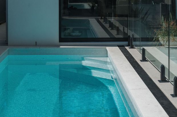 Close-up architectural view of a contemporary swimming pool beside a light paved deck and frameless glass balustrade. The pool has turquoise mosaic-like tiling, integrated submerged steps, and strong reflections of the surrounding modern glazing and architecture.