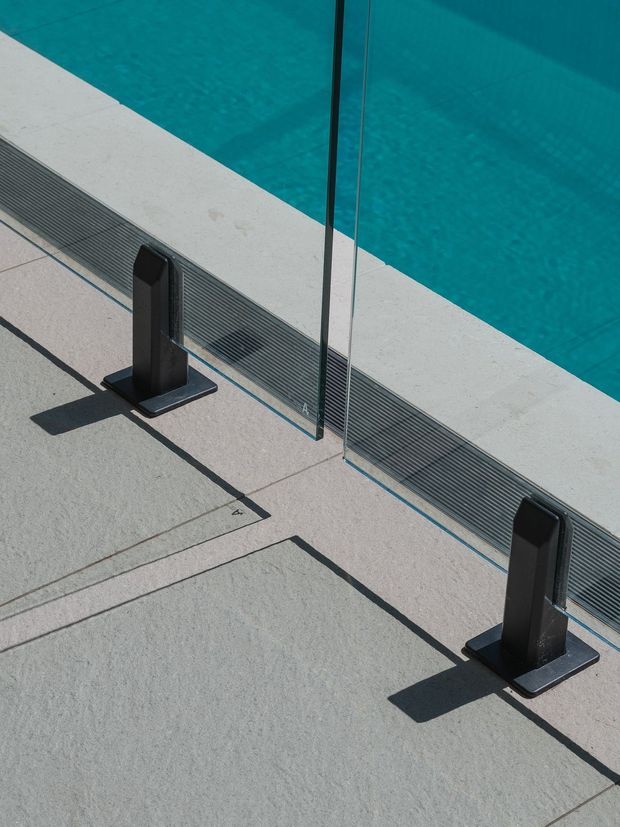 Close-up architectural detail of a swimming pool edge with vivid turquoise water, pale concrete paving, a linear drainage grate, and a frameless glass safety barrier supported by dark metal posts. Strong geometric shadows create a clean, contemporary outdoor composition.