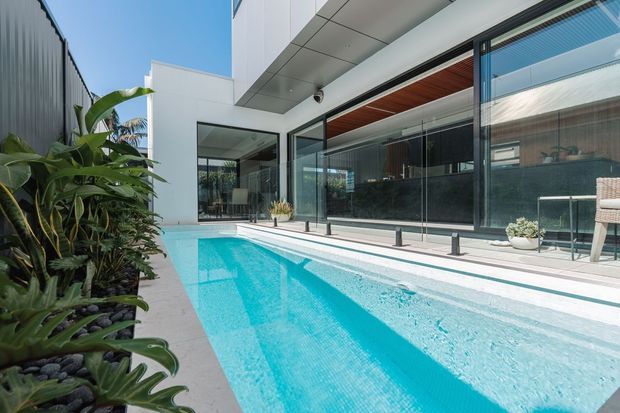 Contemporary residential courtyard with a long turquoise swimming pool beside full-height glass walls. The white rendered facade, projecting flat roof, dark framing, and warm timber soffit create a crisp modern indoor-outdoor connection, complemented by lush tropical planting and a pale paved pool surround.