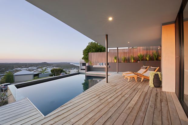 An elevated contemporary outdoor terrace with a timber deck, covered ceiling, infinity-edge swimming pool, sun loungers, built-in planter beds, and a panoramic suburban landscape beyond. The space has warm timber tones, dark structural elements, and a calm open-air atmosphere.