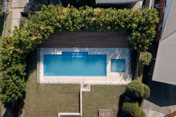 Aerial view of a landscaped residential backyard featuring a rectangular swimming pool with an integrated shallow square section, surrounded by pale paving, a dark timber deck, mature planting, and lawn. Adjacent residential buildings and a paved access path are visible at the edges.