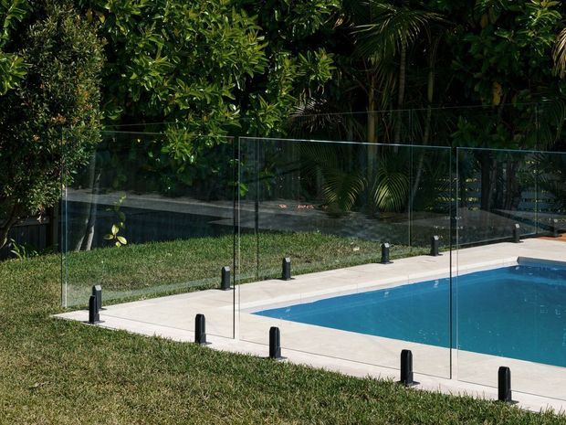A landscaped outdoor swimming pool bordered by pale paving and a frameless glass safety barrier with black posts. Dense tropical greenery forms the backdrop, and the pool water is a vivid blue under bright daylight.