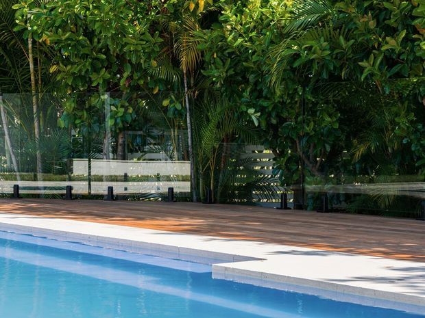 A bright outdoor swimming pool bordered by a pale tiled or concrete pool edge and a broad timber deck. Dense tropical greenery and glass fencing surround the pool, creating a private resort-like setting.