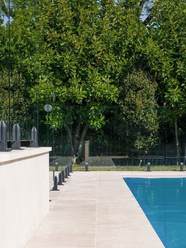 A landscaped outdoor swimming pool area with a broad pale stone-paved surround, clear glass safety fencing, dark metal fence posts, and dense mature greenery screening the perimeter. A simple light-coloured rendered structure or pool feature wall occupies the left foreground.