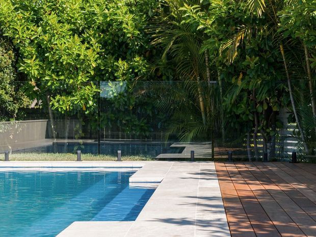 A sunny outdoor swimming pool area surrounded by dense tropical greenery. The pool is bordered by pale stone or concrete paving, with a broad timber deck and glass fencing along the garden edge. Dark-framed glass panels and low bollard lights define the landscaped boundary.
