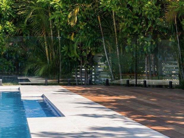 A sunlit swimming pool area surrounded by dense tropical landscaping and a transparent boundary fence. The pool has a crisp white-edged surround, with a broad terracotta-toned paved deck shaded by palms and other lush vegetation.