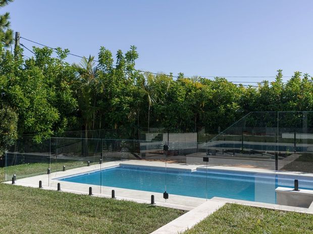 A sunny outdoor swimming pool surrounded by a transparent glass safety fence, with pale concrete paving, trimmed lawn, dense tropical greenery, and a clear blue sky. The image focuses on the pool area and its landscaped setting.