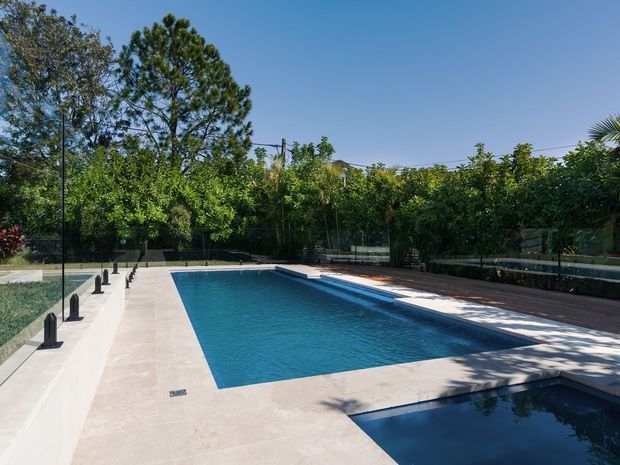 A modern outdoor swimming pool area with a long rectangular main pool and adjoining shallow or spa pool, surrounded by pale stone or concrete paving. Glass balustrades with dark posts line one side, while timber decking, dense greenery, and mature trees create a private landscaped setting.