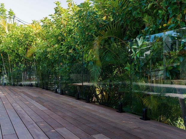A spacious timber deck or terrace bordered by transparent glass balustrades, overlooking dense tropical and subtropical planting. The deck boards are warm brown timber, while the frameless-looking glass panels are supported by dark posts and fittings. The setting is leafy, private, and outdoors.