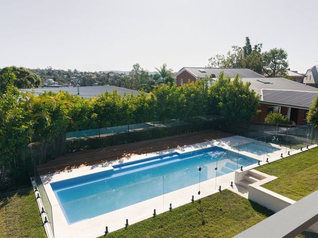 Elevated view of a long rectangular swimming pool enclosed by frameless glass safety fencing, with a pale paved surround, landscaped hedging and lawn. Suburban houses and a waterway or bay are visible beyond the property.