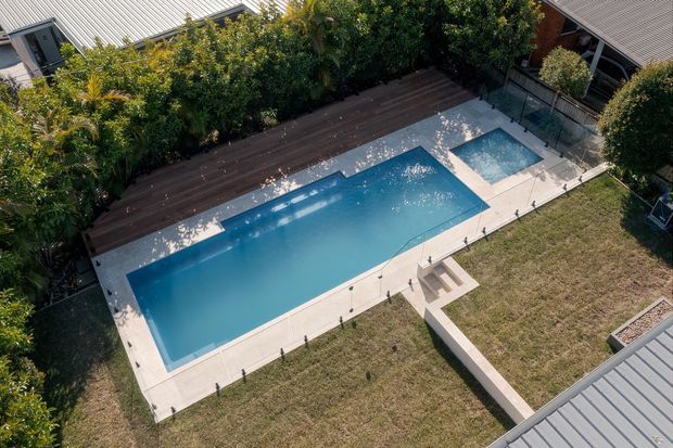 Aerial view of a landscaped residential backyard dominated by a large rectangular swimming pool with a smaller adjoining spa or plunge pool. The pool is surrounded by pale concrete paving, with a raised dark timber deck along one side, dense screening vegetation, lawn, and a narrow connecting walkway.
