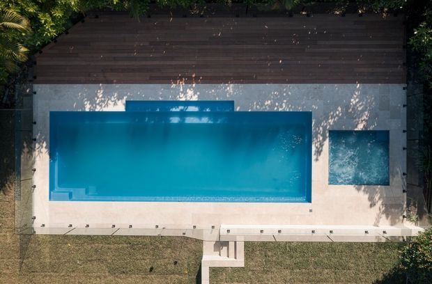 Aerial top-down view of a rectangular swimming pool with an attached square spa, set within a pale paved terrace. A dark timber deck borders the upper edge, with lawn and dense planting surrounding the pool area.