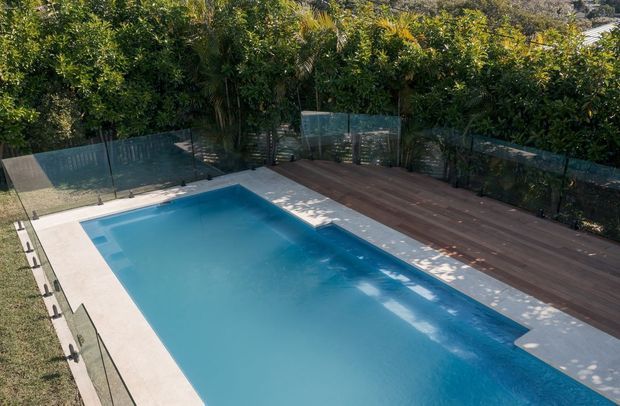 A modern rectangular swimming pool bordered by pale paving and a wide dark timber deck, enclosed with transparent glass safety fencing and surrounded by dense leafy garden planting. The image focuses on the landscaped pool area and outdoor entertaining space.