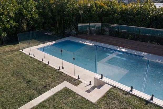 Outdoor swimming pool viewed from above, enclosed by frameless glass safety fencing with a timber deck along the far edge and a lawned garden surrounding the pool.