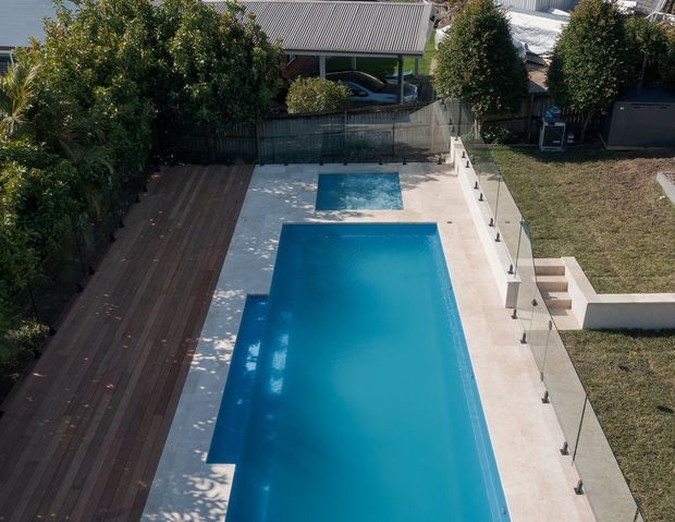 Elevated view of a landscaped residential pool area with a long rectangular swimming pool and smaller square pool or spa, surrounded by pale concrete paving, dark timber decking, glass balustrading, planted screening, and a stepped lawn edge. A neighbouring residential structure and carport are visible beyond the boundary.