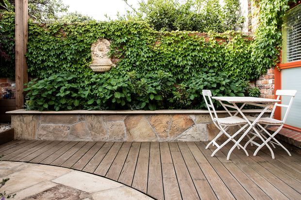 A small, leafy outdoor courtyard or deck with weathered timber decking, a low natural-stone retaining planter wall, dense climbing greenery, and a compact white folding café table and chairs. A timber post and part of a building with horizontal window blinds and orange-red trim are visible at the right edge. The setting feels intimate, garden-like, and residential.