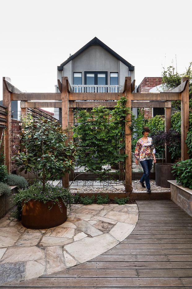 A landscaped urban courtyard garden viewed from a timber deck, featuring a curved natural-stone paved path, raised planters, mature shrubs and climbing greenery. A substantial timber pergola or garden screen frames the space, with a compact gabled dwelling and neighbouring brick buildings visible beyond.
