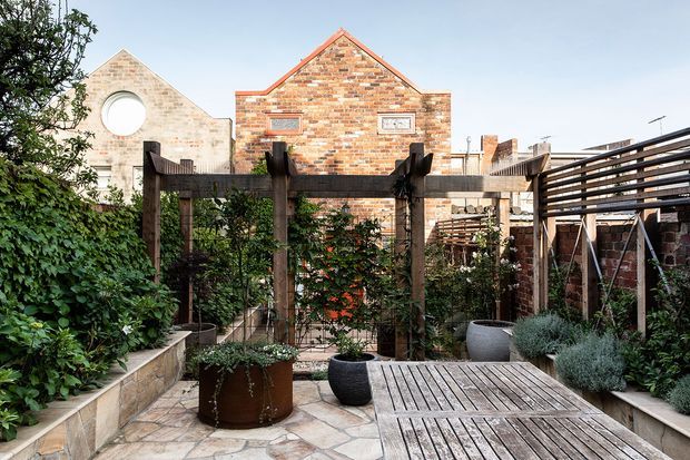 A landscaped urban courtyard garden with raised stone planting beds, timber pergola structures, climbing greenery, potted plants, and a timber deck platform. Brick residential buildings form the backdrop, including a gabled facade and a circular window, creating a private, enclosed outdoor setting.
