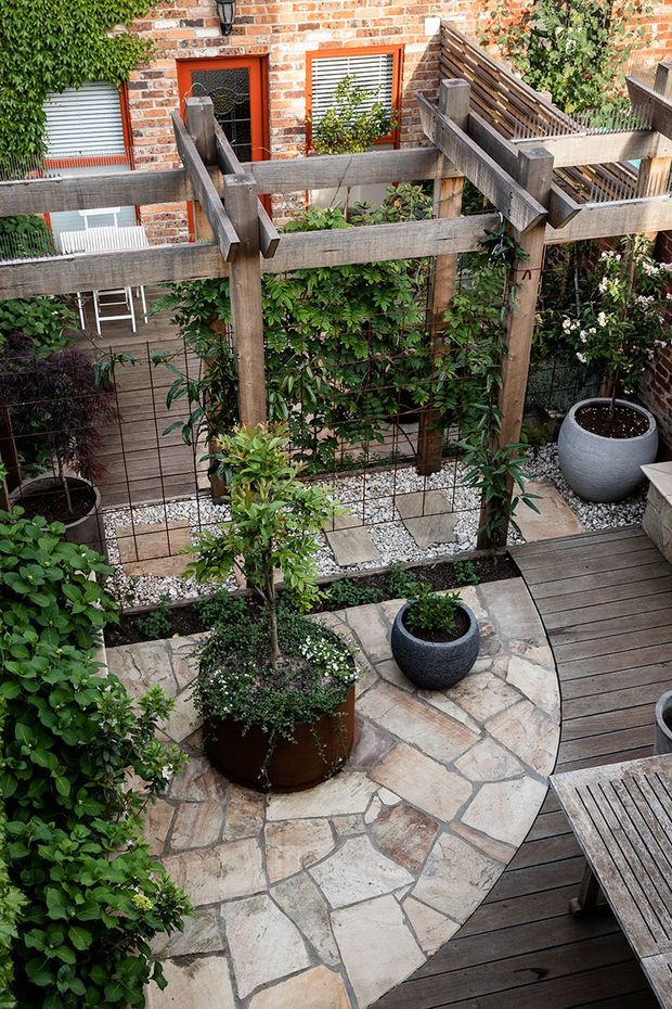 A compact landscaped courtyard viewed from above, featuring a rustic timber pergola and trellis, climbing greenery, raised and potted plants, pale irregular stone paving, white gravel, and dark timber decking. A red brick residential wall with shuttered windows forms the backdrop, creating a layered urban garden setting.