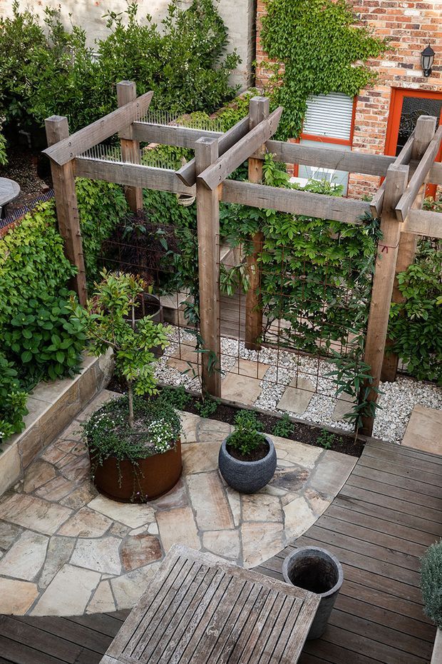A compact landscaped courtyard garden viewed from above, featuring a timber pergola or trellis structure covered with climbing greenery, raised planters, potted plants, irregular stone paving, and a timber deck. A red brick wall with a small window and exterior light forms the backdrop.