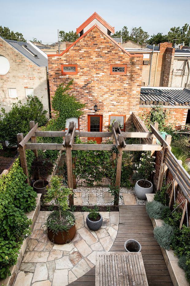 A compact urban courtyard garden viewed from an elevated timber deck, featuring raised timber pergola and railings, dense planting, potted greenery, irregular natural-stone paving, and a narrow timber walkway. Beyond the garden is a red brick building with a prominent gabled roof detail and small decorative windows, surrounded by adjacent urban brick structures.