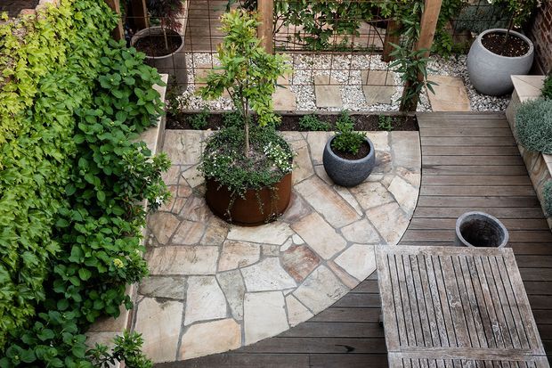 A compact landscaped courtyard viewed from above, featuring an irregular natural-stone paved area bordered by dark timber decking. Dense green planting climbs along the left wall and grows in raised beds and planters, with several large round pots and a timber trellis at the rear. A slatted timber outdoor table occupies the lower right corner, creating a calm, intimate garden setting.