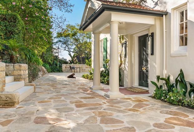 A sunlit traditional-style home entrance with a covered portico supported by classical columns, a dark glazed front door, white rendered walls, and a broad irregular natural-stone paved courtyard or walkway. Lush garden planting, stone steps, low retaining walls, and a terracotta tiled roof edge frame the inviting exterior.