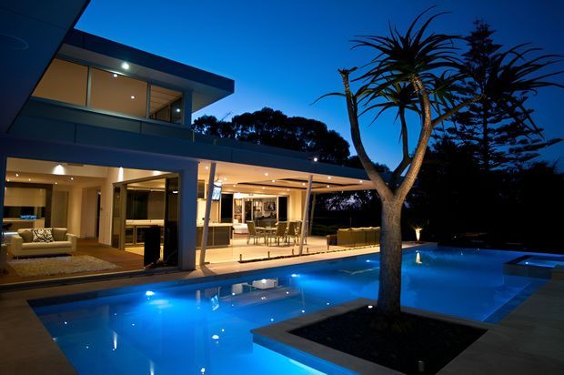 Twilight exterior view of a contemporary multi-level residence with extensive glazed living areas, a covered outdoor entertaining terrace, and a large illuminated swimming pool. A sculptural landscaped tree sits within a raised planter beside the pool, while warm interior lighting contrasts with the deep blue evening sky.