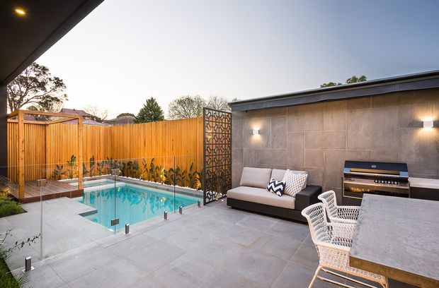 Contemporary outdoor entertaining area with a small swimming pool enclosed by frameless glass fencing, large-format grey tiled paving, timber privacy fencing, and a raised outdoor lounge beneath a roof overhang. A rendered or tiled feature wall includes wall lighting, a built-in barbecue or outdoor kitchen, and a decorative metal privacy screen. Outdoor dining furniture and planting complete the courtyard setting.