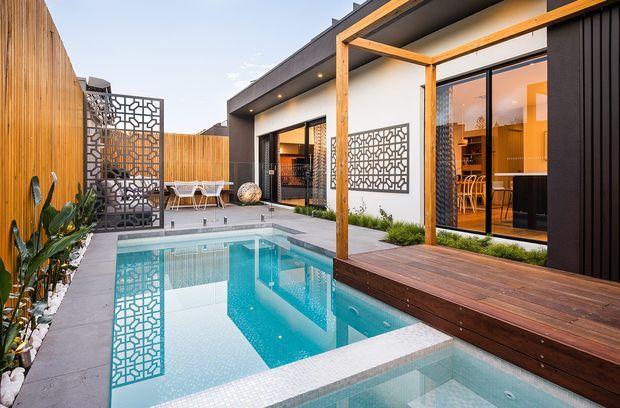 A contemporary courtyard-style residential exterior centered on a compact swimming pool and raised spa. The space features dark vertical cladding, white rendered walls, warm timber framing and decking, patterned metal privacy screens, large glazed openings, and landscaped planting along a tall timber boundary fence. Interior dining and living areas are visible through the glazing.