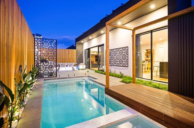 Dusk exterior view of a contemporary single-storey home arranged around a compact courtyard pool. The scene features a timber deck and pergola-like structure, dark vertical cladding, pale rendered walls, large glazed sliding doors, decorative geometric privacy screens, warm recessed lighting, and lush planting beside the turquoise swimming pool and spa.