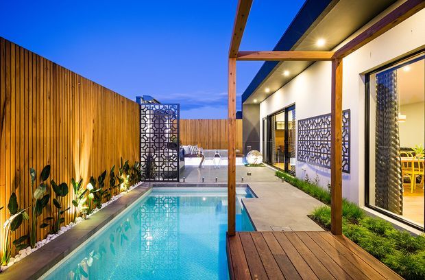 Twilight view of a contemporary courtyard-style home with a narrow rectangular swimming pool, timber decking, warm vertical timber privacy fencing, landscaped planting, decorative geometric screens, and a covered outdoor walkway/pergola. Large glazed openings reveal a bright interior dining area.