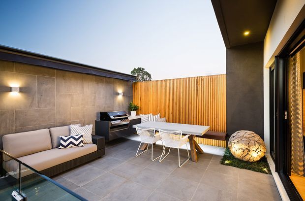 A contemporary covered outdoor courtyard entertaining area with a tiled floor, built-in seating, outdoor dining table and chairs, barbecue, timber slatted privacy screen, concrete-clad wall, glass balustrade, and feature lighting. The space connects directly to the interior through large glazed folding or sliding doors.
