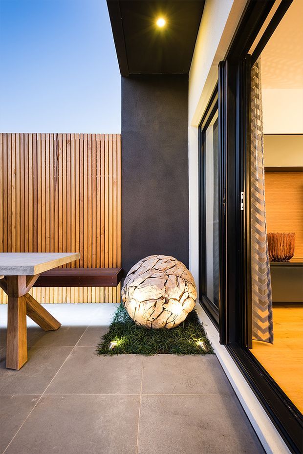 A contemporary outdoor courtyard or patio beside a home, featuring large grey tiled paving, a timber dining table and bench, vertical timber privacy fencing, and a sculptural stone feature set into a small planted bed with uplighting. Black-framed sliding glass doors open into a warmly lit interior, with textured masonry detailing visible around the opening.
