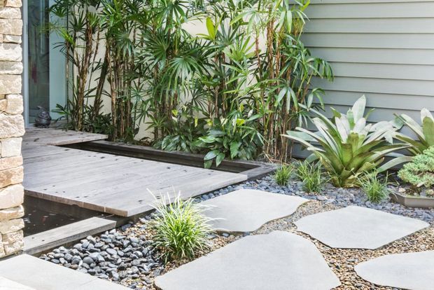A landscaped courtyard or garden pathway beside a contemporary building, featuring large irregular concrete stepping slabs set in small gravel and smooth river stones, a raised timber deck and bridge-like walkway, dense tropical planting, and horizontal grey exterior cladding. A pale stone wall frames the left edge.