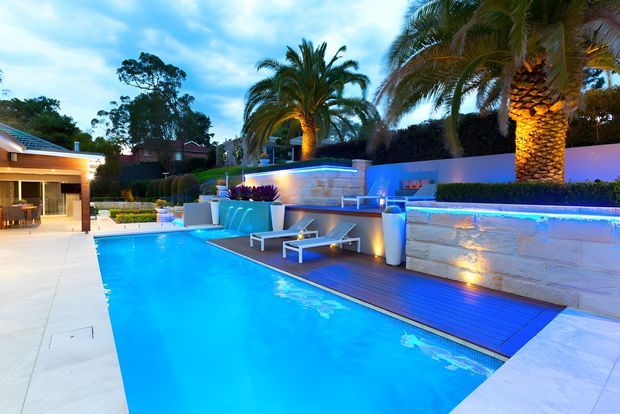 A luxurious landscaped backyard pool area at dusk, featuring a large rectangular swimming pool, pale stone paving and retaining walls, timber decking, integrated blue LED lighting, sun loungers, tropical palm trees, and a covered outdoor entertaining structure with a kitchen or bar area.